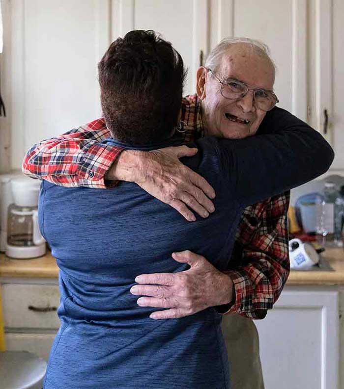 An older gentleman hugs a Meals on Wheels volunteer in his kitchen