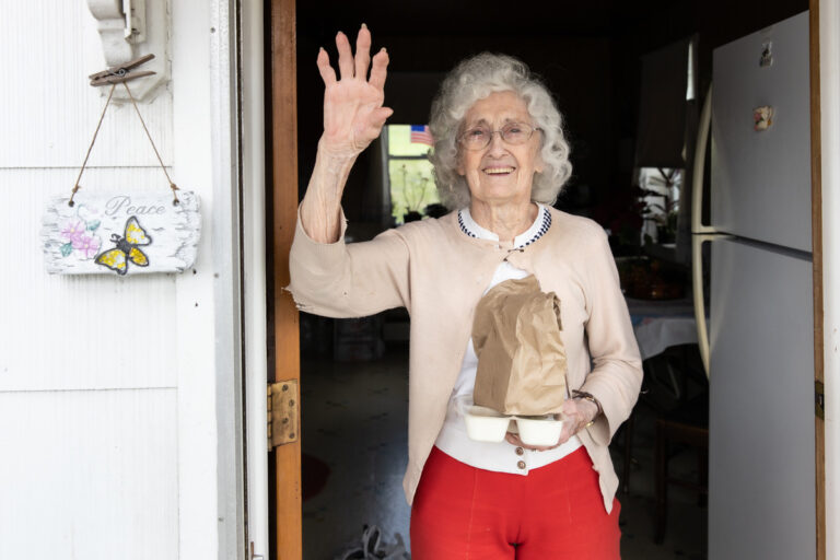Smiling older woman wave from her doorstep while holding a delivered meal, representing the warmth, indpendence, and joy Meals on Wheels brings to older adults living at home.