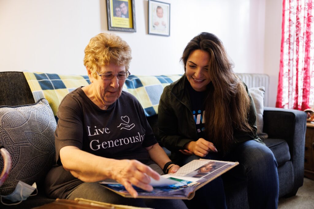 An older adult and a Meals on Wheels volutneer sit together on a couch looking through a photo album, sharing a quiet, joyful moment that reflects compaionship, trusts, and meaningful connection during an in-home visit