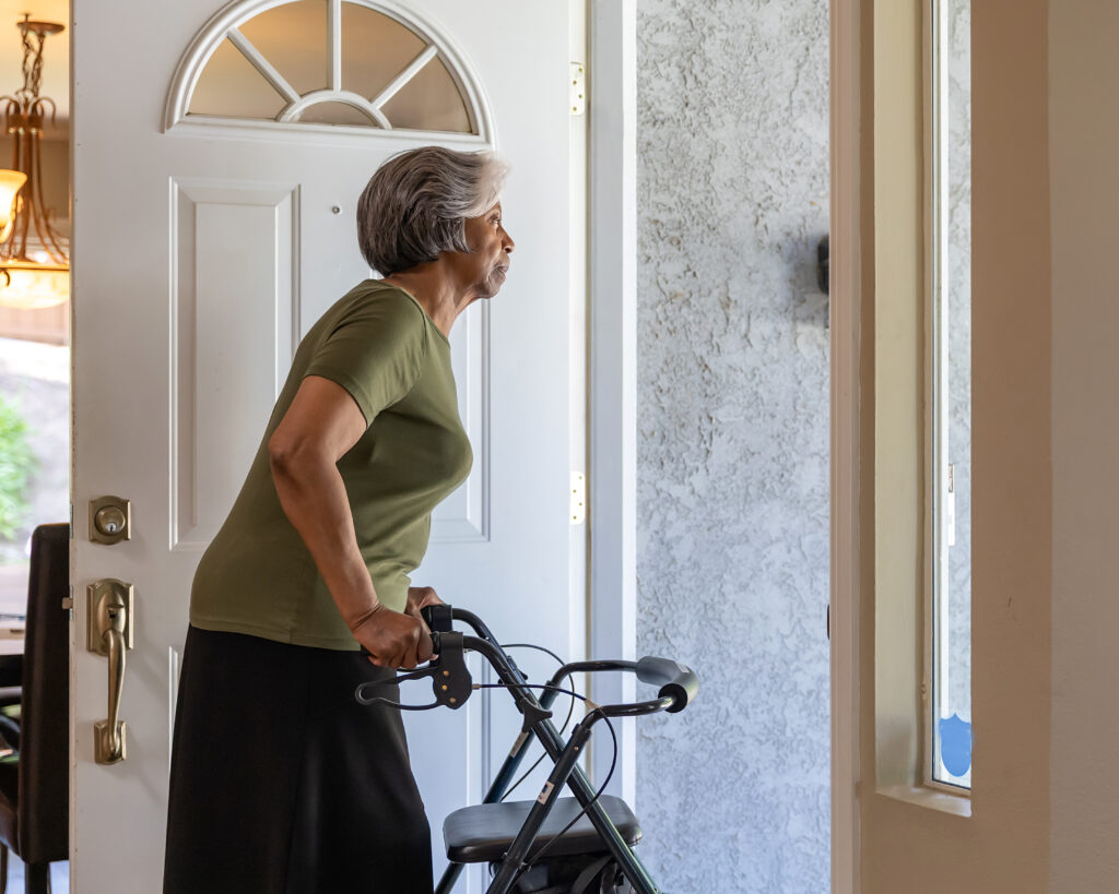 Older adult using a walker stands at the front door of her home, looking outside as she waits to greet a visitor or meal delivery.