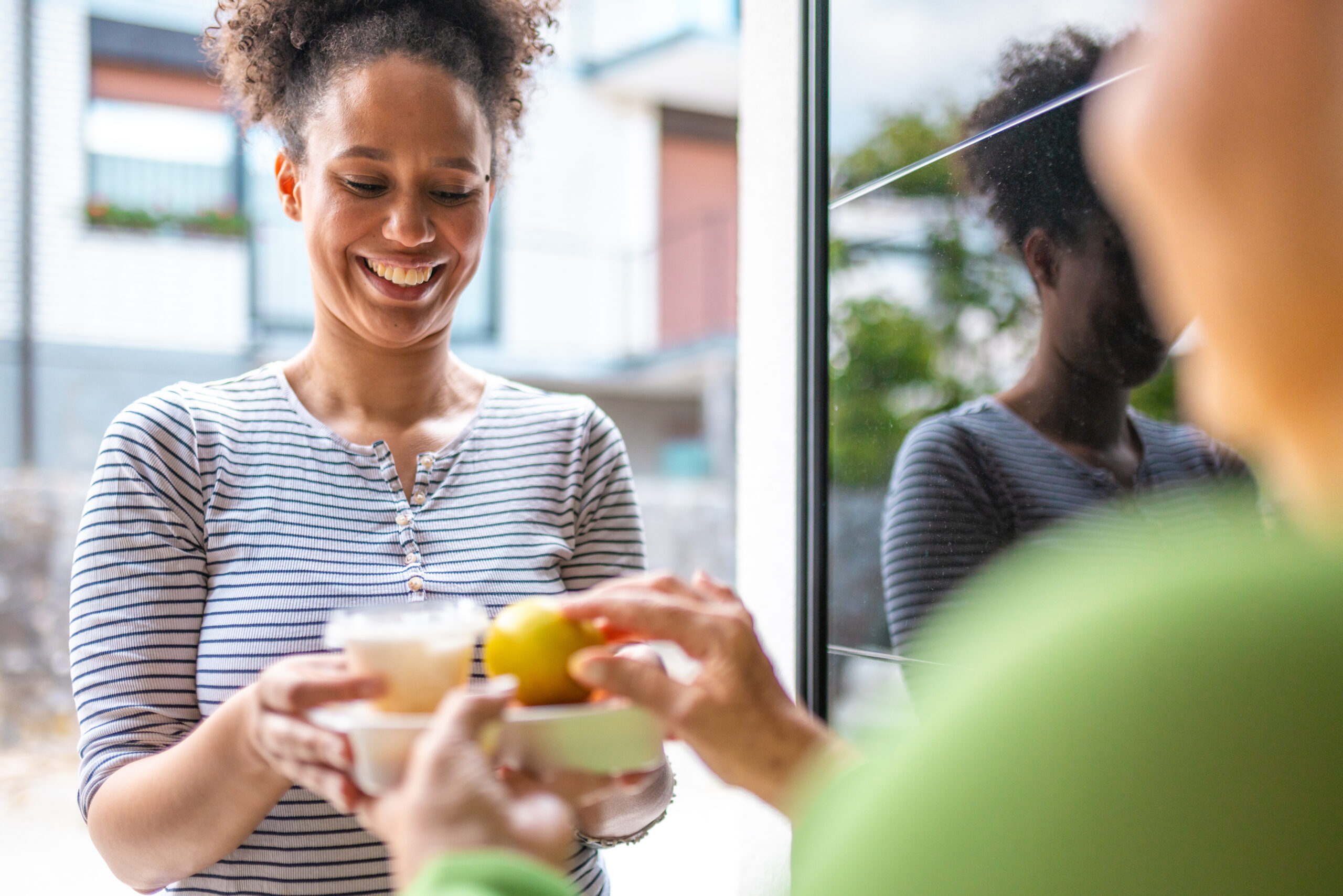 A smiling Meals on Wheels volunteer hands a meal tray through a glass door to an older adult, capturing a warm moment of connection during a home meal delivery.