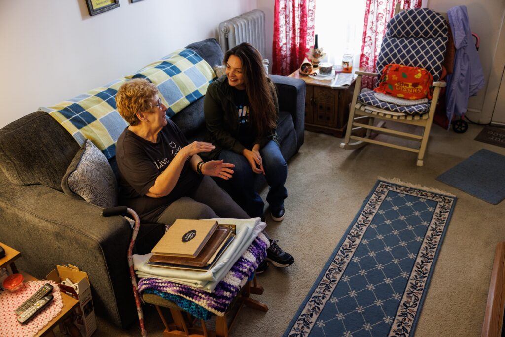 A Meals on Wheels volunteer sits on a couch talking with an older adult in her living room, sharing a warm, attentive conversation that highlights companionship and connection during an in-home visit