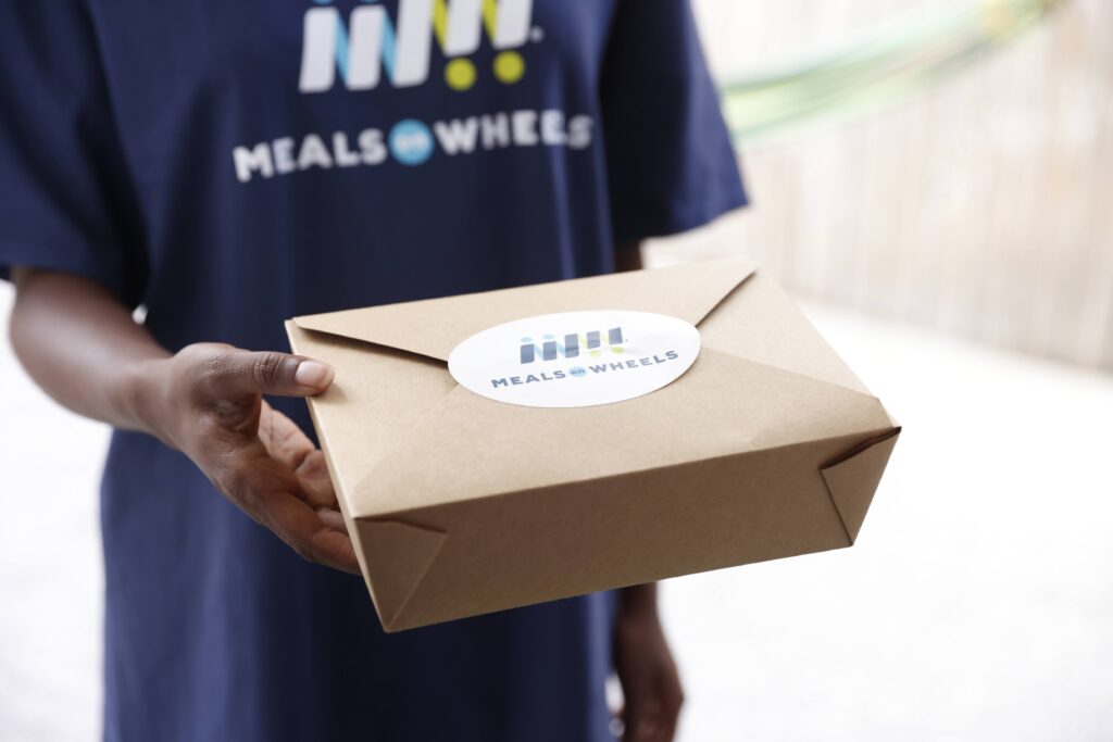 Meals on Wheels volunteer wearing a navy shirt hands a brown meal delivery box labeled with Meals on Wheels logo.