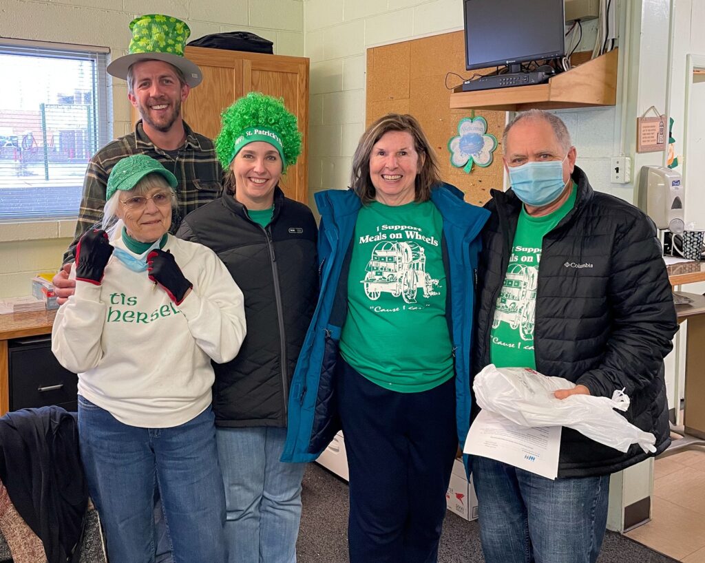 Group of Meals on Wheels volunteers in Greeley, Colorado smiling and wearing green St. Patrick's Day outfits and shirts that say "I Support Meals on Wheels," gathered indoors before delivering meals.