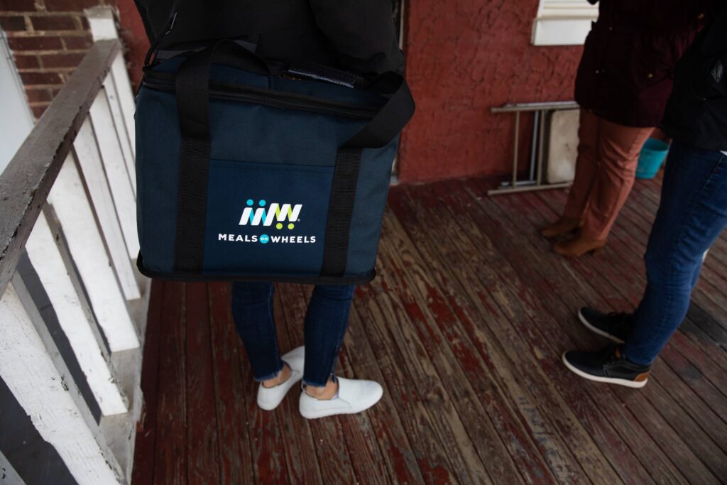 Volunteer carries an insulated Meals on Wheels delivery bag onto a front porch where another person waits at the door.