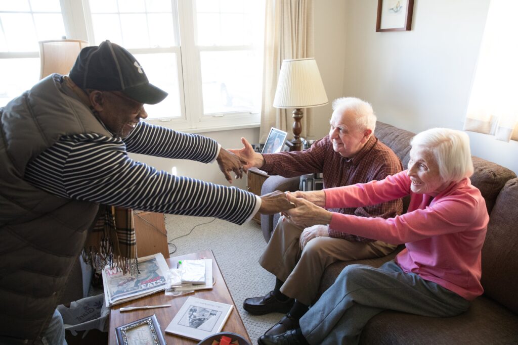 Smiling visitor reaches out to clasp hands with an aging couple seated on their living room couch during a home visit.